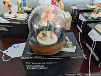 Miniature ceramic figurine of a barefoot boy in straw hat under clear dome with wooden base, sitting on original box.