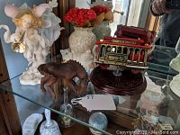 Photo showing white porcelain fairy with trumpet, white cat vase with flower details, wooden horse figurine, and red San Francisco cable car model on wooden base on glass shelf.