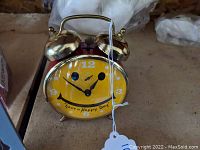 Vintage metal twin bell alarm clock with yellow happy face dial and 'Have a Happy Day' text, placed on brown surface, with a white tag attached.