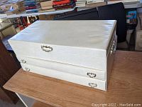 White jewelry box with a smooth finish, showing the top and front sides with metal handles and clasps on a wooden table.