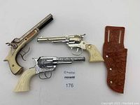 Three cap gun toys alongside a brown suede holster with floral embossing, all placed on white background.
