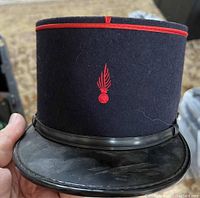 Front view of dark blue French kepi hat with red piping and embroidered flaming grenade insignia. Black leather visor visible.