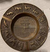 Top view of brass ashtray showing Chinese zodiac animal reliefs around rim and engraved pavilion center, identifying Pan Mun Jon Korean War truce talks site.