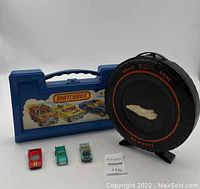 Photo showing three toy cars, two carrying cases including a blue plastic Matchbox case and a black Rally Hot Wheels tire-shaped case