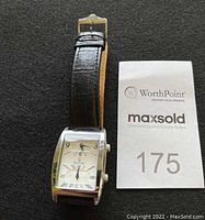 Men's Bulova dual time watch with black leather strap, resting on black fabric background. Included auction or inventory card with number 175.