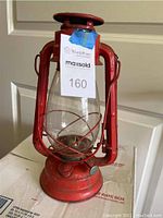 Full view of red metal kerosene lamp with clear glass globe and metal protective frame, labeled with a lot number tag.