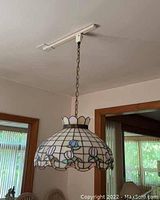 Tiffany style ceiling light fixture hanging from chain attached to ceiling track mount, showing overall view in a dining area with chairs and windows.