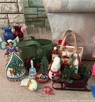Photo showing a range of ceramic and fabric Christmas decorations including a ceramic angel figurine, red sled planter with greenery, small snowman, fabric tote with plush ornaments, glass vases, and glass bell ornaments.