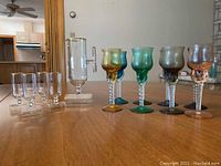 Photo showing the entire set of one pitcher, four clear cordials with gold rims, and eight colored twisted stem cordials arranged in a row on a wooden surface.