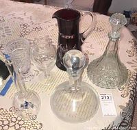 Overview of entire set on lace tablecloth: red cut glass pitcher, clear cut glass decanter with stopper, clear glass decanter with round stopper, and two clear glass vases.