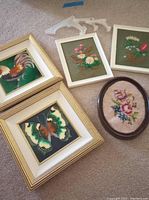 Photo showing five framed pieces: two gold-framed decorative tiles with butterfly and rooster designs, two smaller rectangular floral embroidery pieces in white frames, and one oval framed floral embroidery needing repair.