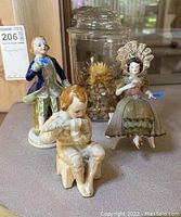 Three Occupied Japan porcelain figurines including a man in blue coat, seated child with instrument, woman in bonnet with fan, next to a broken glass jar with lid.