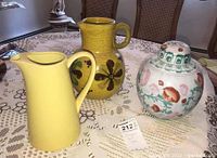 Photo showing three ceramic items: yellow pitcher, decorated pottery pitcher, and ceramic ginger jar with lid on lace tablecloth.