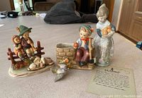 Photo showing all four figurines together on a countertop: Lladro girl with oranges, Erich Stauffer boy with ax, bird, and Gobel two children with dog.