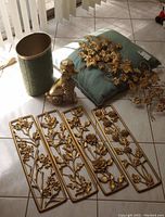 Photo showing four gold metal floral wall panels, gold metal poodle figurine, green pillow with gold floral decoration, and green wastebasket on tile floor with sunlight.