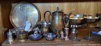 Shelving display with multiple silverplate and pewter items including large round tray, covered dish, pitchers, goblets, salt shaker, pepper grinder, and footed bowls. Items show tarnish and patina.