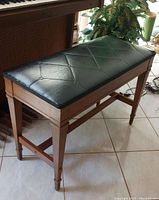 Perspective angled photo of wooden piano bench showing dark green vinyl cushioned seat and wooden legs with supports.