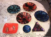 Wide shot showing five acrylic plates with major abstract paint patterns (red, blue, green, black) and one bright coral vintage ceramic ashtray with finger rests.