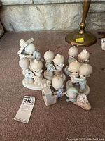 Photo showing a group of Precious Moments figurines, including children and an angel figure, arranged on carpeted floor with a brass lamp in background.