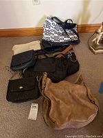 Eight assorted handbags and purses arranged on a carpet near a wall and wooden baseboard, showing various sizes and styles including large tan, woven black, navy/white tote, and others.