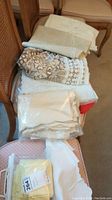 Stack of assorted folded table cloths and napkins on a chair, some appear lace or woven pattern, neutral colors