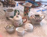 Seven ceramic items displayed on lace tablecloth including teapots, vases, cream and sugar set
