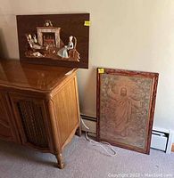 Two religious wall art pieces, one lacquered wood relief on top of a cabinet and one framed religious print leaning against wall