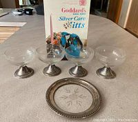 Four weighted sterling silver base footed glasses, glass plate with sterling silver rim and Goddard Silver Care polishing mitts box displayed on table.