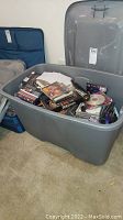 Large gray plastic storage bin filled with assorted DVD movies and music CDs scattered inside on carpet.