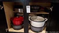 Open cabinet showing red and grey enameled cast iron pots stacked on left and white and grey enameled cast iron pots stacked on right, with black bundt pan on upper center shelf