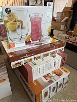 Stacked boxes showing Copper Chef Pro 8-piece pan set, Tramontina 3-piece pan set, and Libbey Ellington 16-piece glassware set with pink drink and lemon garnish on the packaging.