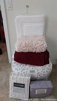 Stack of bedding and throws on a small chair including king floral comforter, two throws, and packaging for bath mats and sheets.