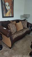 Full view of love seat with pillows featuring dark brown leather and patterned fabric upholstery under a framed painting.