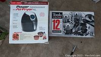 Photo showing boxed Power Air Fryer XL 5.3 QT 1700W unit alongside boxed Tools of the Trade 12-piece stainless steel cookware set on carpeted floor.