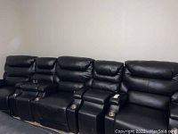 Front overview showing a row of four black reclining theater chairs with two console units between them; stainless steel cup holders visible.