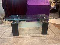 Front side angled view of rectangular coffee table with clear beveled glass top, black wooden legs, metal support rods, and frosted glass bottom shelf on a light carpeted floor
