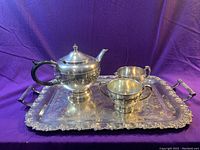 Tea set displayed on purple background with all pieces visible showing engraved details and black handle on teapot