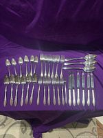 Full layout of all flatware pieces laid out on purple fabric background showing spoons, forks, knives, and serving pieces.