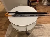 Two large black umbrellas on white round table, one with wooden handle and sleeve showing Doorman logo, the other black handled.