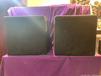 Pair of black upholstered square footstools shown from the outside, lit by natural indoor light with a purple cloth backdrop.