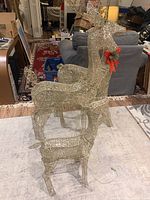 Full view of three wireframe reindeer lawn ornaments of varying sizes arranged indoors on a rug. Largest reindeer has a red bow attached at the neck, all show the clear light strings wrapped around metal frames.