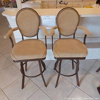 Front view of two matching heavy metal bar stools with beige fabric upholstery on seats, arms and round backs, set on a tiled floor in front of a kitchen counter.