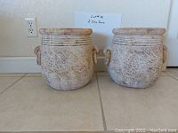 Pair of decorative clay pots with grapevine relief pattern and handles, placed on a tiled floor.