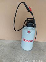 Front view of white 2-gallon Gilmour sprayer showing handle, black lid, hose, and red brand name on tank.