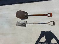 Two shovels laid out on concrete surface: one sand shovel with rust on blade and wooden handle, and one flat tip shovel with black metal handle and metal blade.
