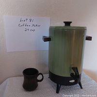 Photo showing a 24 cup coffee maker with green cylindrical body, black spigot and base on table, with brown coffee cup.
