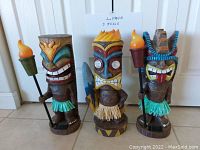 Photo of three distinct painted wooden tiki idol statues each around 18 inches tall, with torches and vibrant paint detail.