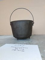 Front view of the black cast iron kettle showing its 3 legs and hanging handle, placed on a floor with a visible handwritten note stating lot details.
