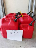 Photo showing seven 5-gallon red plastic gasoline containers and one 2-gallon red plastic gasoline container. Each features a black flexible pouring spout with a yellow cap.