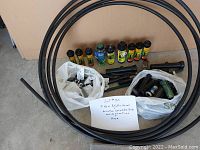 Top-down photo showing coiled black hose tubing arranged in a circle, yellow and black sprinkler rotator heads lined up, black plastic pipes, two white bags filled with assorted sprinkler parts and connection fittings, and a handwritten note describing the lot.